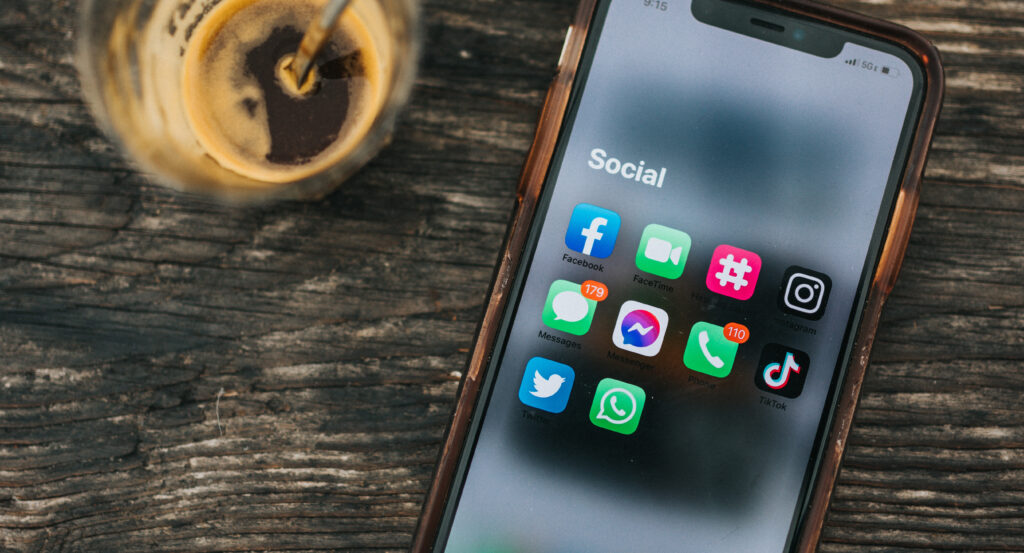 Smartphone mit einem Glas Kaffee nebenan. Auf den Bildschirm sind verschiedene Social-Media Apps zusehen, darunter Facebook, Instagram, Twitter und TikTok.