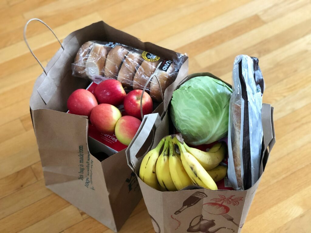 Es ist sind zwei Einkaufstaschen mit Obst und Gemüse zu sehen. Man sieht Brötchen, Äpfel, Bananen und ein Stück Kopfkohl.